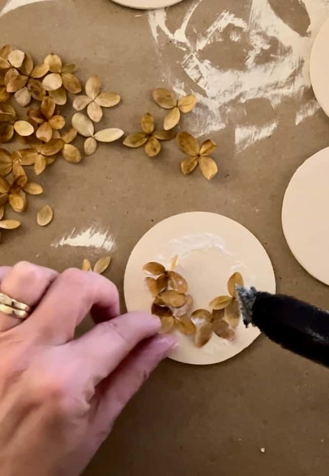 A hand uses a hot glue gun to attach small dried flowers onto a round beige circle on a craft table. Additional dried flowers and other circles are scattered around the workspace.