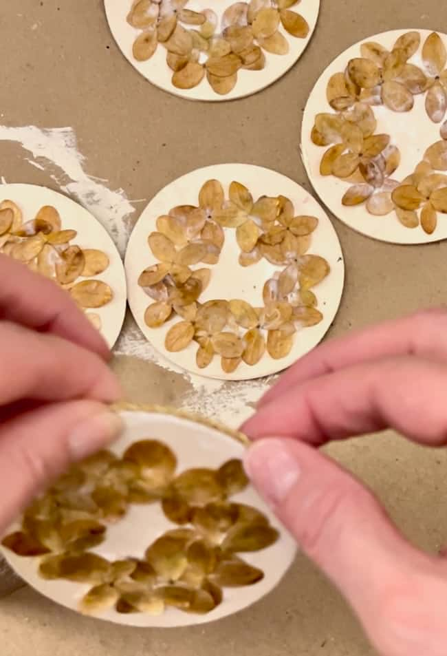 Hands holding a circular ornament made of seeds arranged in a flower pattern. Several similar seed crafts are placed on a brown surface in the background.