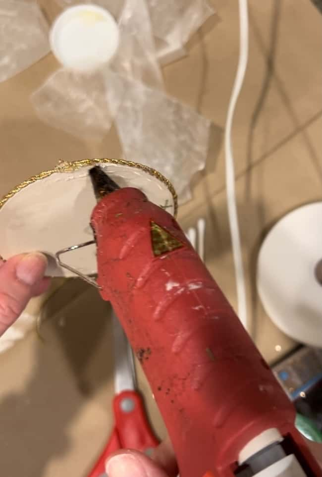 A person is using a red hot glue gun to attach a round, gold-trimmed object to a translucent material. Scissors and a few crafting materials are visible on the table.