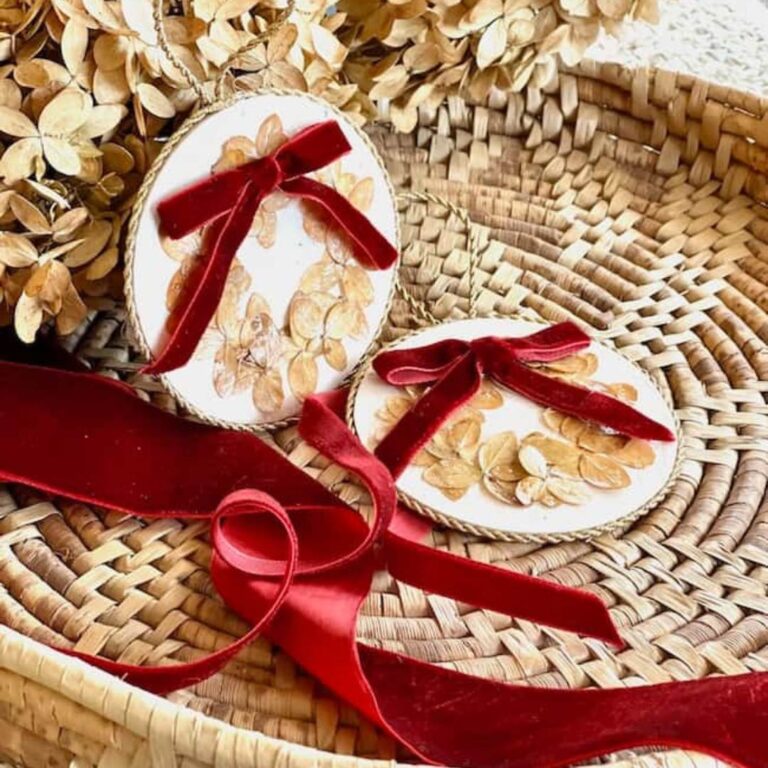 Two round ornaments decorated with dried petals and red velvet bows are placed on a wicker tray, embodying DIY creativity. Nearby, a red velvet ribbon is laid out. Dried flowers surround the ornaments, adding a rustic touch to these hydrangea Christmas treasures.