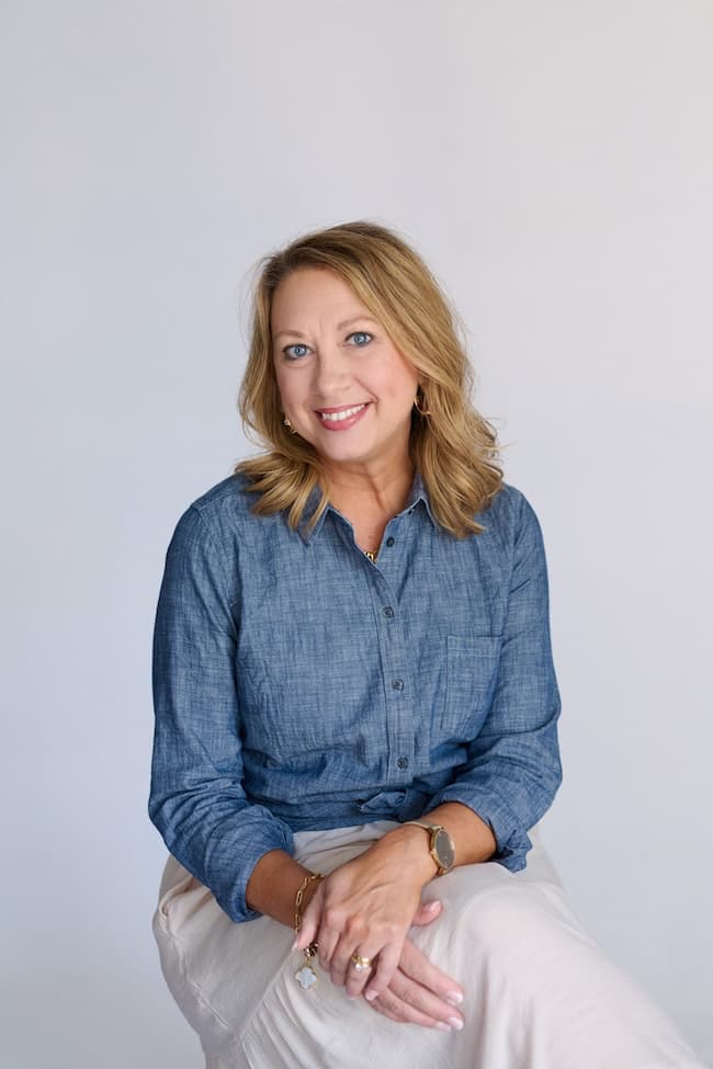 A smiling person with shoulder-length blonde hair, dressed in a denim button-up shirt and beige pants, is seated comfortably at home against a plain, light background. Their hands rest gently in their lap.