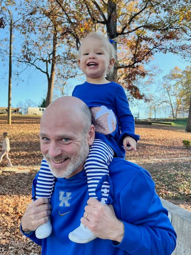 A bald man in a blue hoodie smiles as he carries a smiling toddler on his shoulders, enjoying the crisp air of 11/23/24. The toddler, wearing a blue outfit with striped pants, giggles amid the trees and fallen leaves on this clear day—a perfect moment for Seven on Saturday.