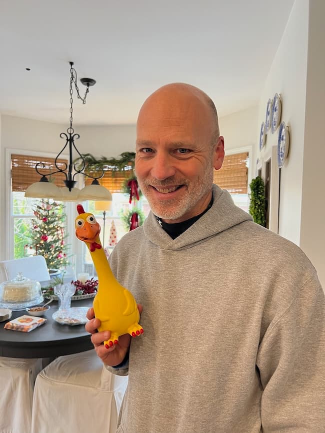 A smiling person in a gray hoodie holds a rubber chicken toy in a festively decorated room, featuring a chandelier, round table, and Christmas garlands. It's as if they're celebrating "Seven on Saturday" or perhaps gearing up for the upcoming 11/30/24 festivities.