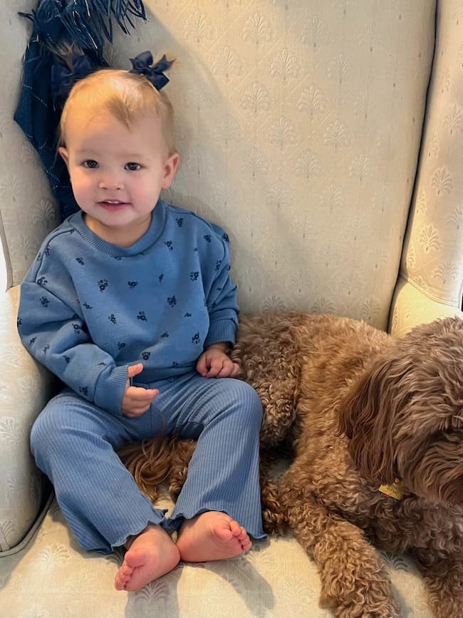 A toddler in a blue outfit sits on an armchair next to a brown dog, like they're enjoying the perfect Sunday moment. With a small bow in their hair, the child smiles at the camera, and the dog rests calmly beside them as if it's 11/10/24—a day of precious memories captured forever.