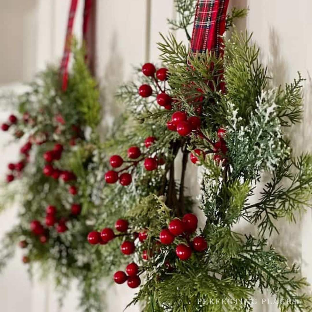 Close-up of two festive wreaths with green foliage, red berries, and frosted tips. Each is hung with a plaid ribbon on a light-colored door, perfect for adding charm to your Seven on Saturday decor blog.