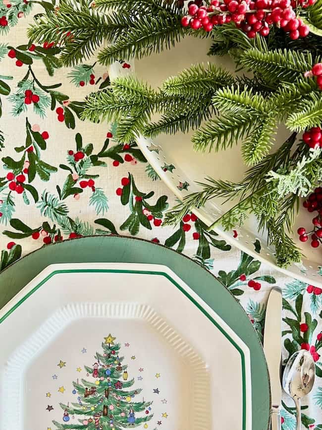 A festive table setting with a plate featuring a decorated Christmas tree design, surrounded by green and red holly-patterned tablecloth. A sprig of evergreen and red berries adorn the top part of the image. Silverware is placed beside the plate.