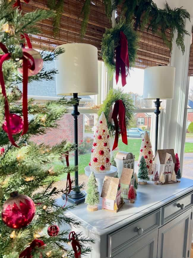 A holiday-themed room with a Christmas tree to the left, decorated with red ornaments. A side table holds miniature houses, trees, and white cone-shaped decorations. Two lamps with white shades stand behind, near a window with greenery and red ribbons.