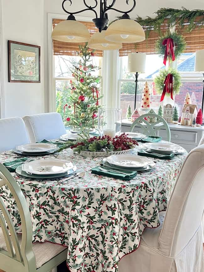 A festive dining room decorated for Christmas with a table set for four. The tablecloth features holly patterns, and the centerpiece includes greenery and berries. A small decorated tree and holiday ornaments are visible by the window.