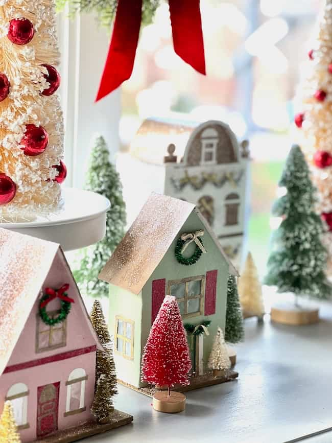 A festive holiday scene with decorated miniature houses and small pine trees adorned with snow. Red and green wreaths decorate the houses, and there's a large red ribbon in the background. The image conveys a cozy, wintry atmosphere.