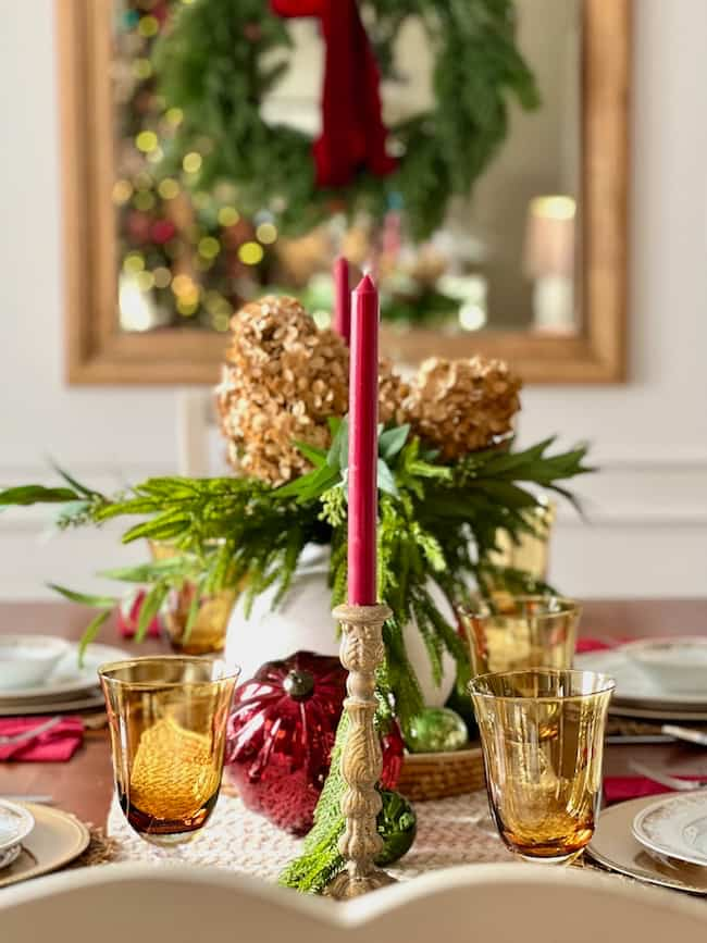 A festive table setting with amber glasses, a centerpiece of greenery and dried flowers, red candles, and decorative ornaments. A wreath is reflected in the mirror above the table, enhancing the holiday atmosphere.