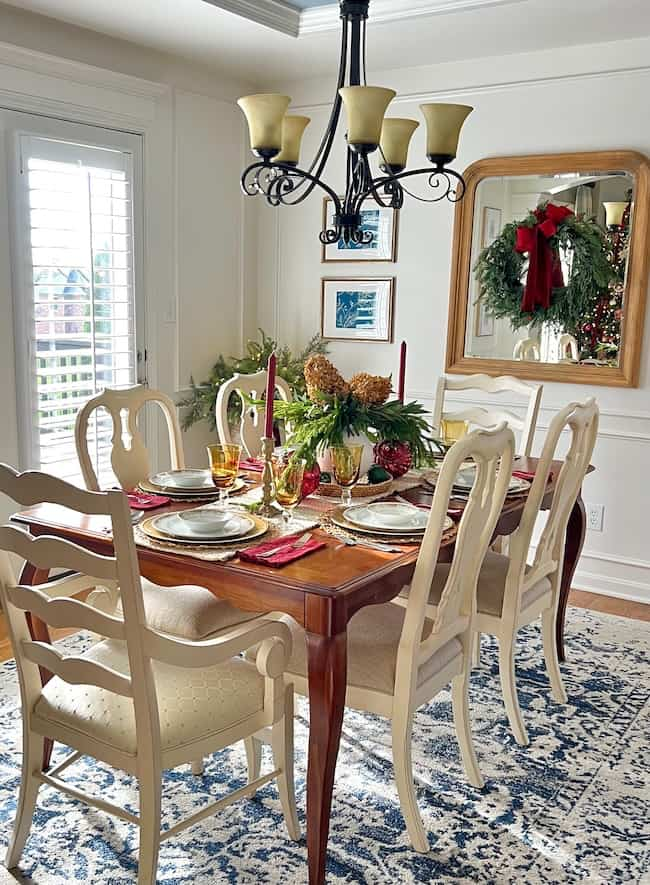 A dining room with a wooden table set for a festive meal. It features red and gold decorations, candles, and a centerpiece. Six white chairs surround the table. A mirror with a wreath hangs on the wall, and a chandelier is above. Natural light enters the room.