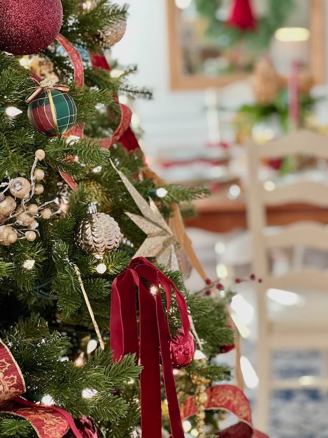 A close-up of a decorated Christmas tree with red and plaid ornaments exudes classic Christmas charm, adorned with ribbons and lights. In the background, a blurred table setting hints at timeless elegance and a festive holiday atmosphere worthy of any delightful home tour.