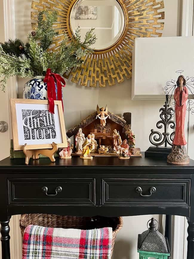 A cozy Christmas display on a black table, featuring a nativity scene, framed art, a potted plant with festive ribbons, and decorative items. A round, ornate mirror hangs above, reflecting the warm, holiday ambiance.