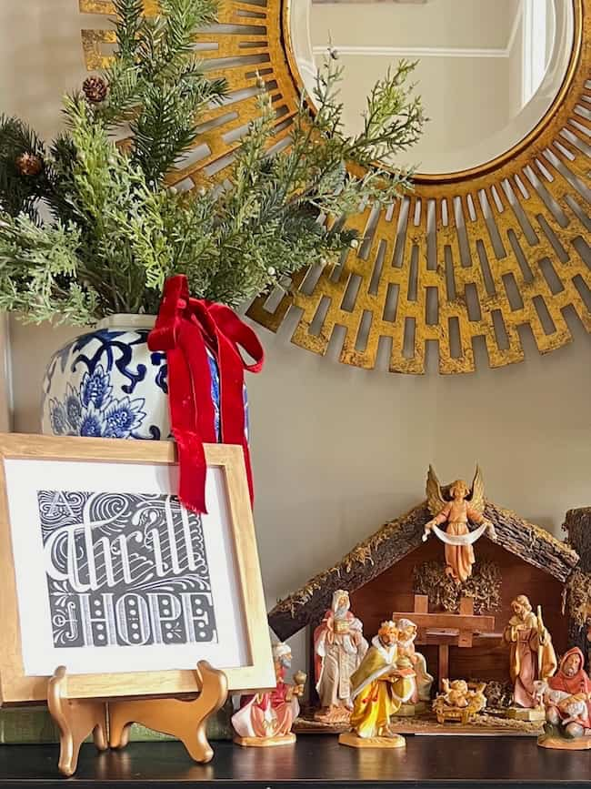 A festive holiday display featuring a gold sunburst mirror, a vase with greenery and a red ribbon, a framed artwork with the word "Hope," and a nativity scene with figurines arranged around a manger under a wooden structure.