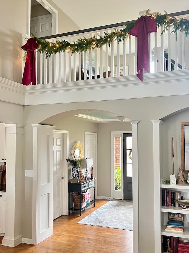 The two-story home interior exudes timeless elegance, showcasing a decorated staircase with a classic Christmas touch. A garland with red bows adorns the upper railing. The hallway features hardwood flooring, a console table with decor items, and a rug near the front door. Shelves are partially visible on the right.