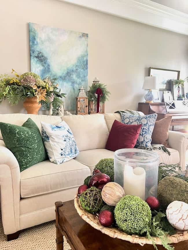 A cozy living room with a beige sofa adorned with green, blue, and brown cushions. A vibrant abstract painting hangs above. The coffee table features a decorative tray with candles and greenery. Lanterns and plants decorate the area.