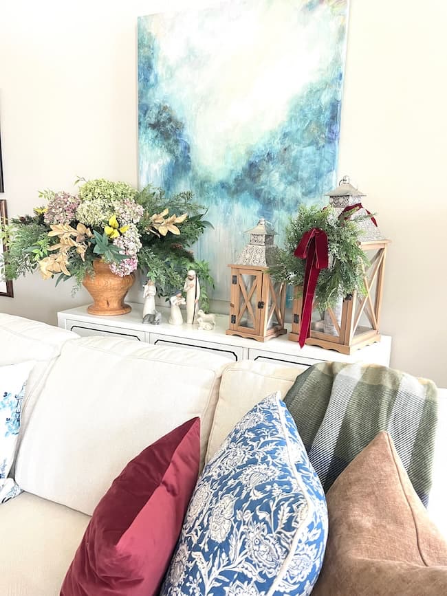 A cozy living room scene with a white couch adorned with colorful pillows exudes timeless elegance. A console table behind it features a large floral arrangement, wooden lanterns with ribbon, and a decorative statue. A blue abstract painting hangs on the wall, completing this elegant home tour.