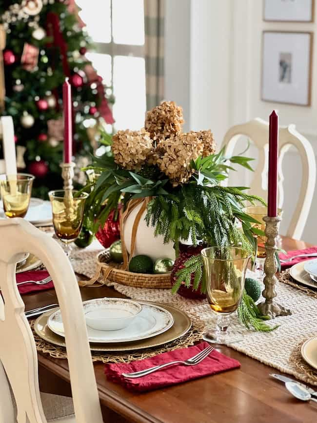 A festive dining table is set with elegant plates, gold goblets, red napkins, and tall red candles. A centerpiece of greenery and dried flowers adorns the table, with a decorated Christmas tree visible in the background.