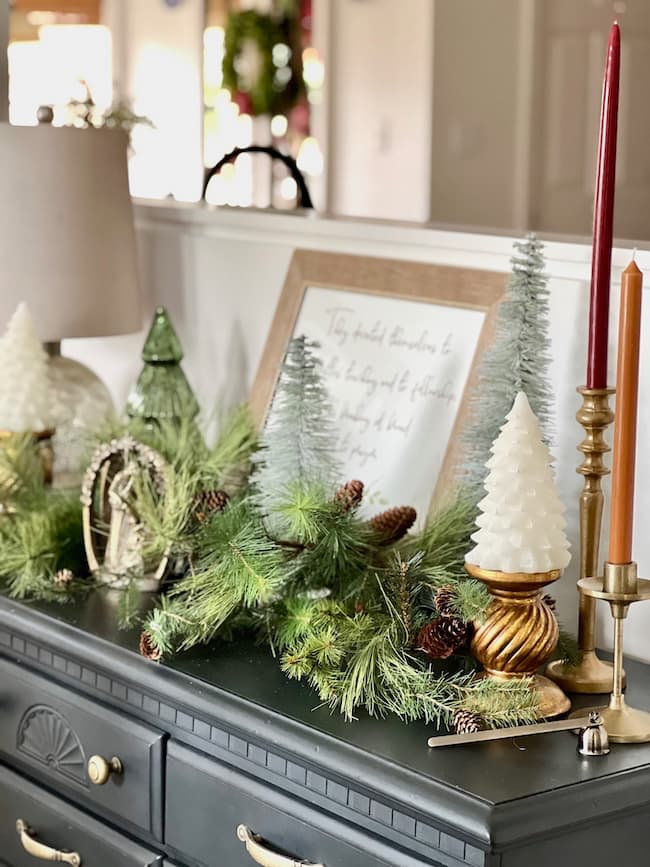 A festive holiday scene on a dark dresser exudes timeless elegance with small pine trees, white tree-shaped candles, and rustic pine cones surrounding a framed quote. Classic Christmas charm is added by a lamp and tall orange and brown candles in the arrangement.