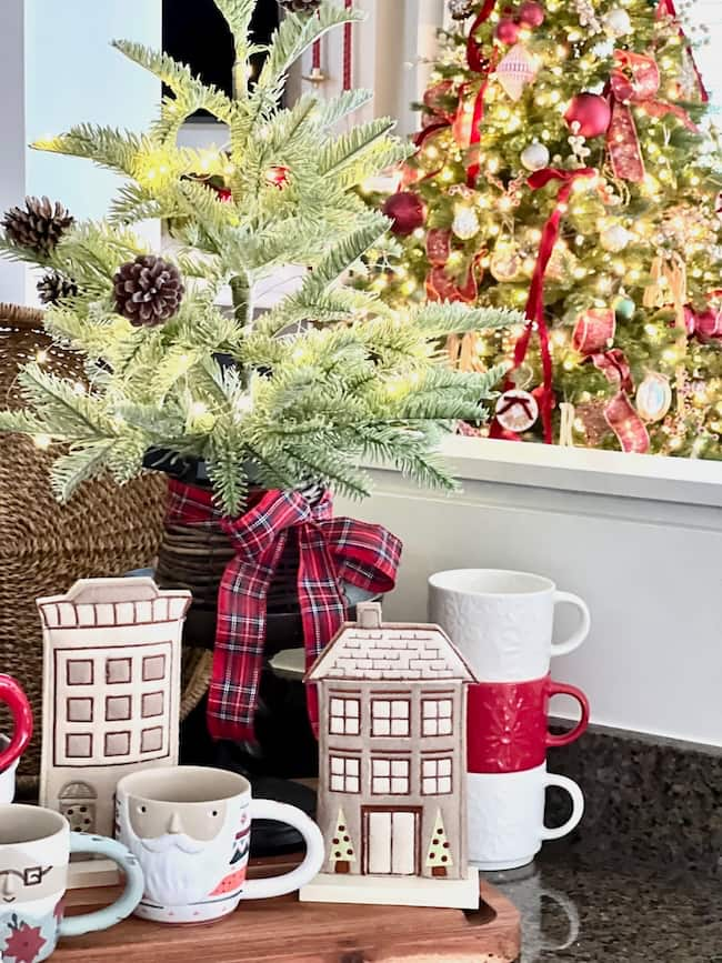 A cozy holiday scene features a small decorated Christmas tree with pinecones and a red plaid ribbon. Nearby, two festive mugs sit beside stacked mugs. In this classic setting, a glowing larger Christmas tree with lights and red ribbons completes the home tour ambiance.