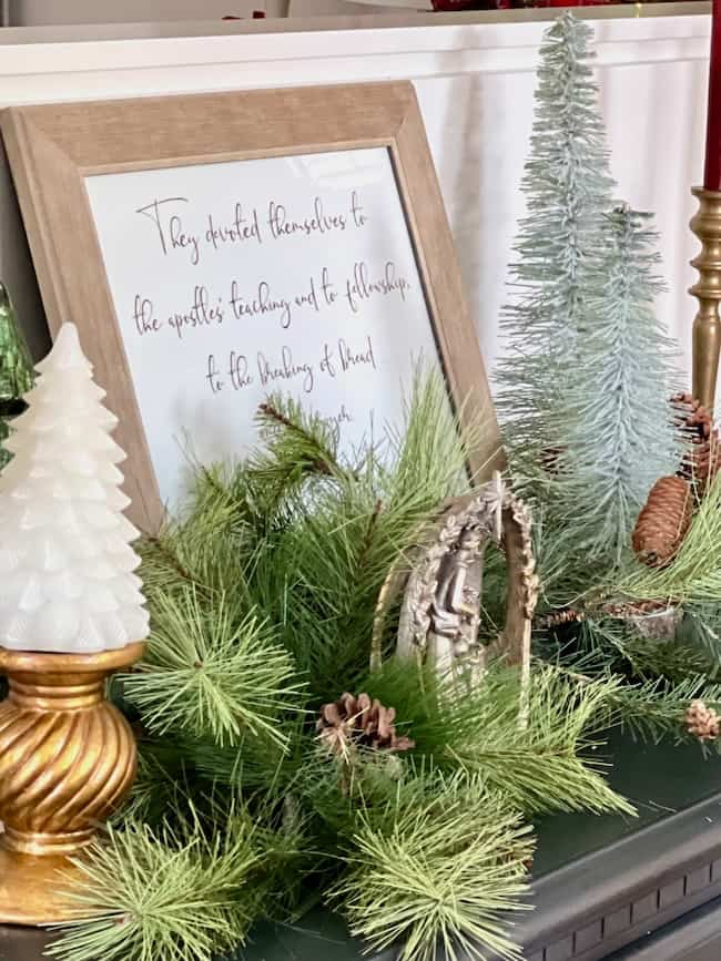 A festive holiday display featuring pine branches, a small nativity scene, a white Christmas tree candle, a framed quote, and miniature evergreen trees. The arrangement includes pine cones and is adorned with winter-themed decor.