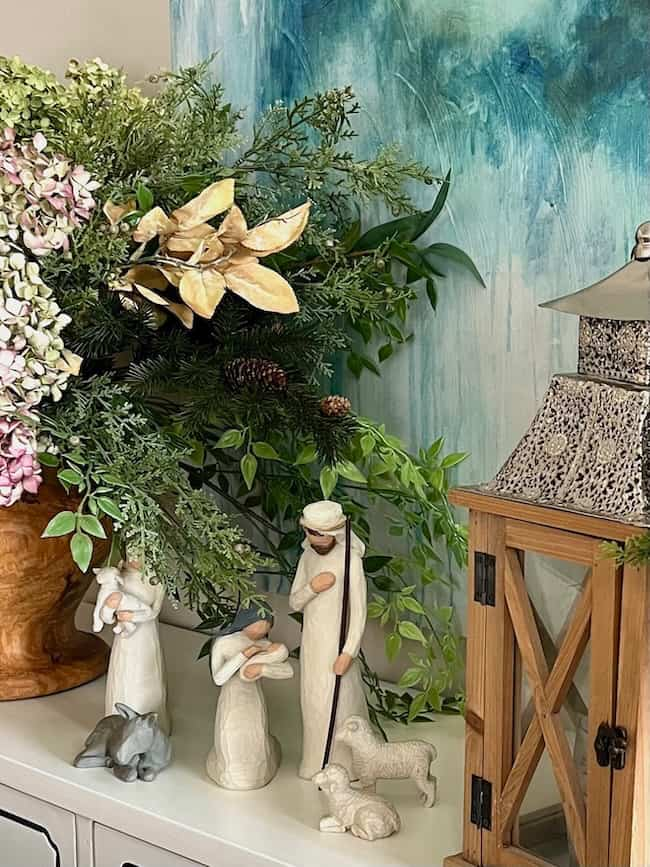 A decorative nativity scene featuring three figurines in white robes, a baby in a crib, a donkey, and a sheep is displayed on a table. Surrounding the scene are lush green leaves and flowers, next to a rustic wooden lantern.