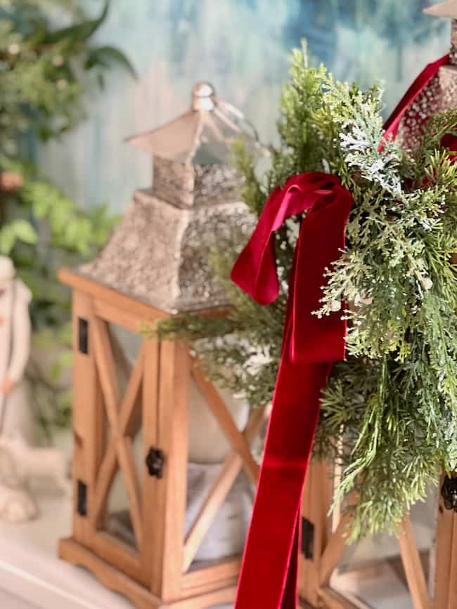 A festive arrangement with a lush green wreath adorned with a red ribbon, placed near two decorative wooden lanterns with candles inside. The background shows a blurred, nature-inspired painting.