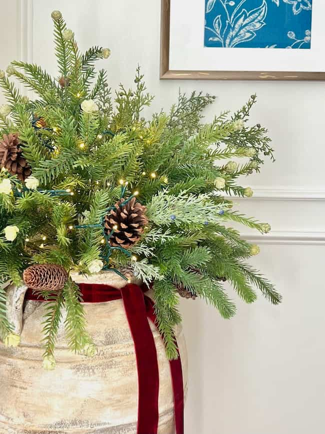 A festive arrangement in a rustic wooden basket filled with evergreen branches, pinecones, and fairy lights. A red velvet ribbon decorates the basket. A framed art piece with blue floral patterns hangs on the wall in the background.