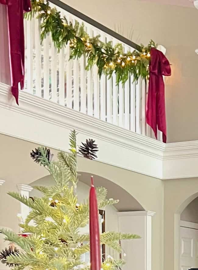 A decorated indoor staircase with a garland of green leaves and twinkling lights. Red bows adorn the railing. In the foreground, a partial view of a Christmas tree with red candles and pinecones adds to the festive atmosphere.