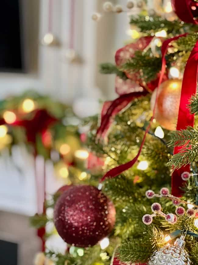 Close-up of a festive Christmas tree decorated with red ribbons, glittery ornaments, and twinkling lights. A blurred background reveals more decorations, creating a warm, holiday ambiance.