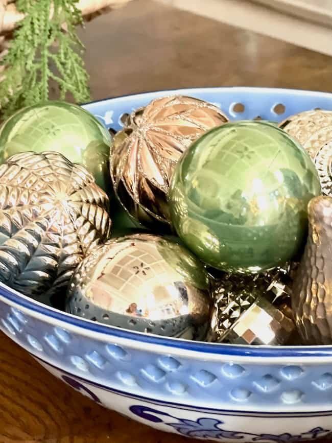 A blue bowl filled with shiny, textured ornaments in green and metallic gold sits on a table. A small evergreen branch is visible in the upper left corner.