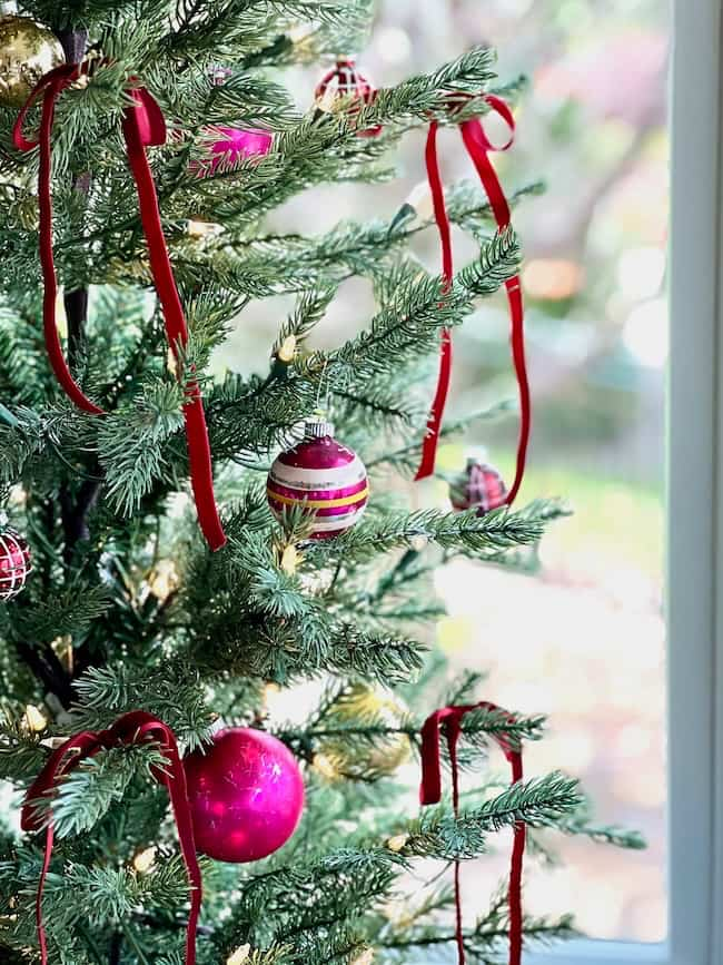 A close-up of a decorated Christmas tree with bright green branches. Red and pink ornaments and red ribbons are neatly hung throughout the tree. Soft natural light filters in through a window in the background.