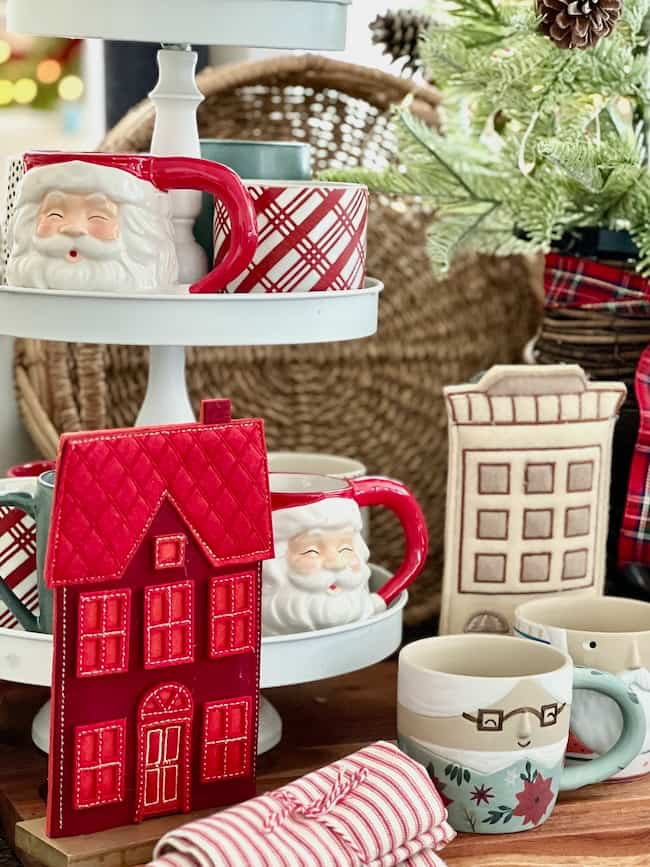 A festive display features tiered shelving with Santa-themed mugs, plaid mugs, and patterned mugs. A miniature red house decoration and a napkin with red stripes are visible alongside Christmas tree branches.