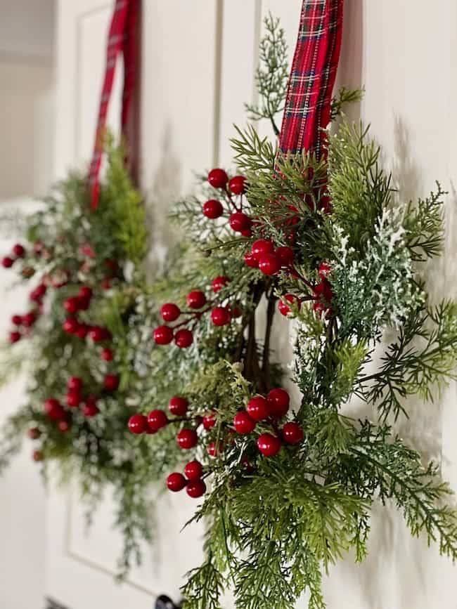 Close-up of two festive wreaths hanging on a door. Each wreath features lush greenery, red berries, and a snowy touch, suspended by red tartan ribbons.