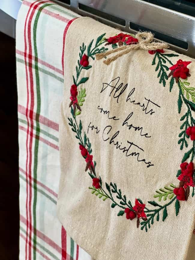 A holiday-themed dish towel hanging on an oven handle features a wreath with red berries. The text inside the wreath reads "All hearts come home for Christmas." A plaid towel is visible in the background.