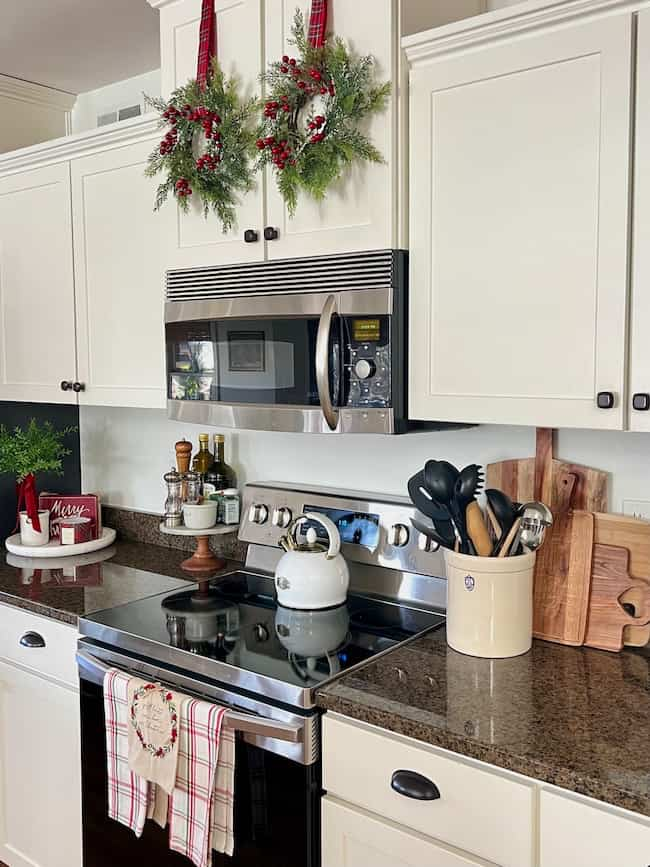 A cozy kitchen decorated for the holidays features white cabinets adorned with wreaths, a stainless steel microwave, a stovetop with a kettle, and a countertop holding utensils, cutting boards, and festive décor.