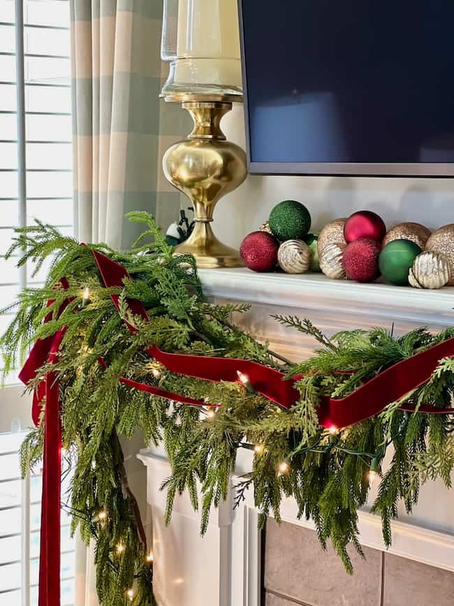 A festive mantel decorated with green garland, red velvet ribbon, and string lights. A collection of red, green, and gold ornaments rests beside a gold lamp. A TV is mounted above, and a window with striped curtains is partially visible.