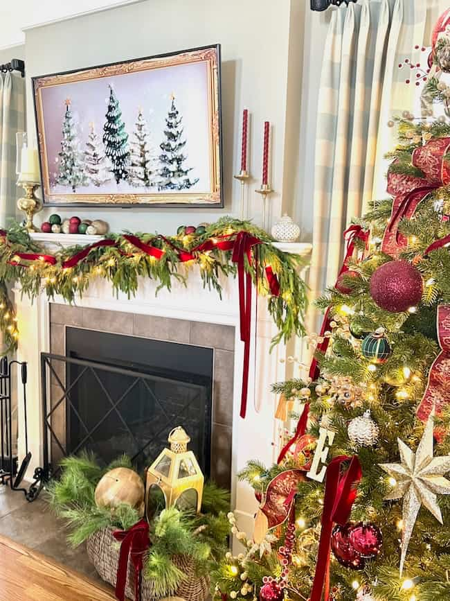 A festive living room decorated for Christmas, featuring a fireplace adorned with greenery and red ribbons, a painting of snowy trees above the mantel, and a Christmas tree with ornaments and lights. A wicker basket with pine decor sits nearby.