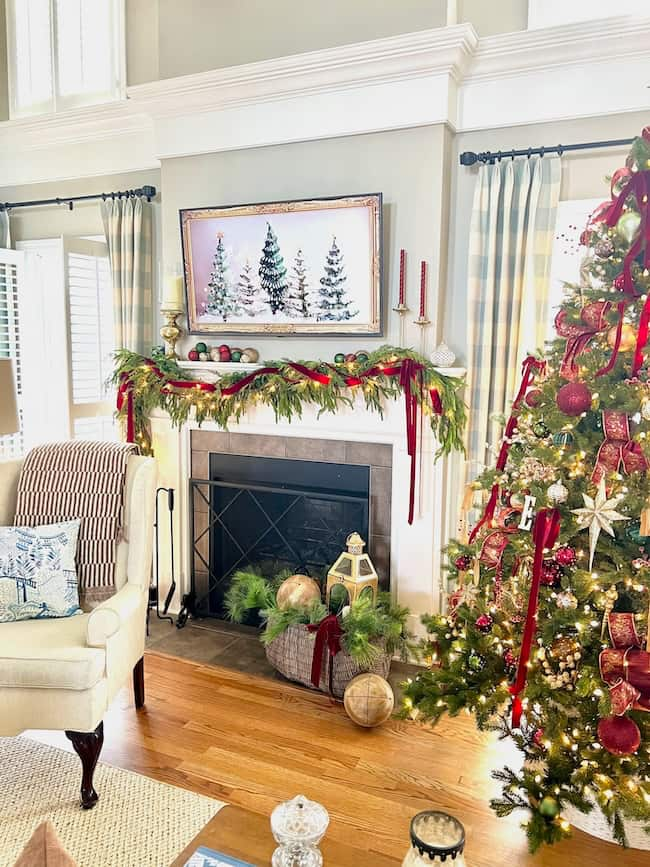 A cozy living room exudes timeless elegance with its Christmas decor. The fireplace is adorned with garland, red ribbons, and candles, capturing a classic Christmas charm. Nearby, a lit tree dazzles in red and gold ornaments as a snowy landscape hangs above the mantel.