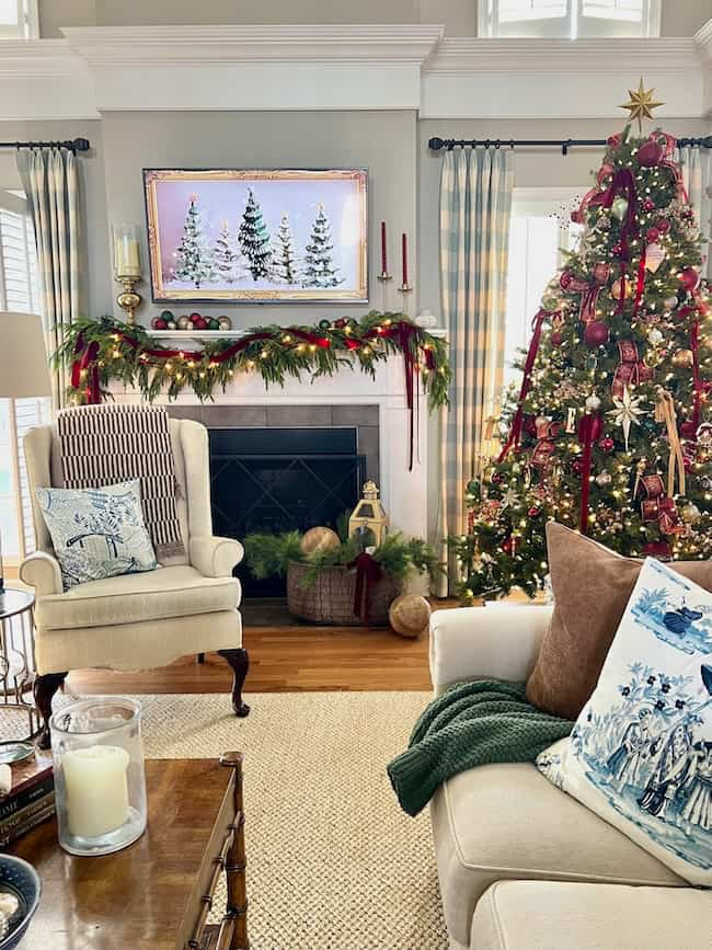 A cozy living room decorated for Christmas with a large tree adorned with lights and ornaments. The fireplace mantel is garnished with garland and red candles. A framed painting of snowy trees hangs above the fireplace. Plush chairs enhance the warmth.