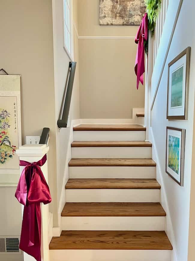 A wooden staircase with light-colored walls, adorned with vibrant pink bows tied to the railing. Framed artwork hangs on the walls, and greenery is visible at the top. Natural light streams in from an unseen window above.