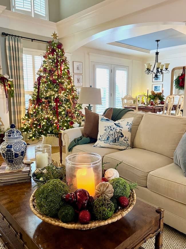 A cozy living room decorated for Christmas with a brightly lit tree adorned with ornaments. A comfortable beige sofa features festive pillows. The coffee table holds a decorative bowl with candles and greenery. Natural light streams in through large windows.