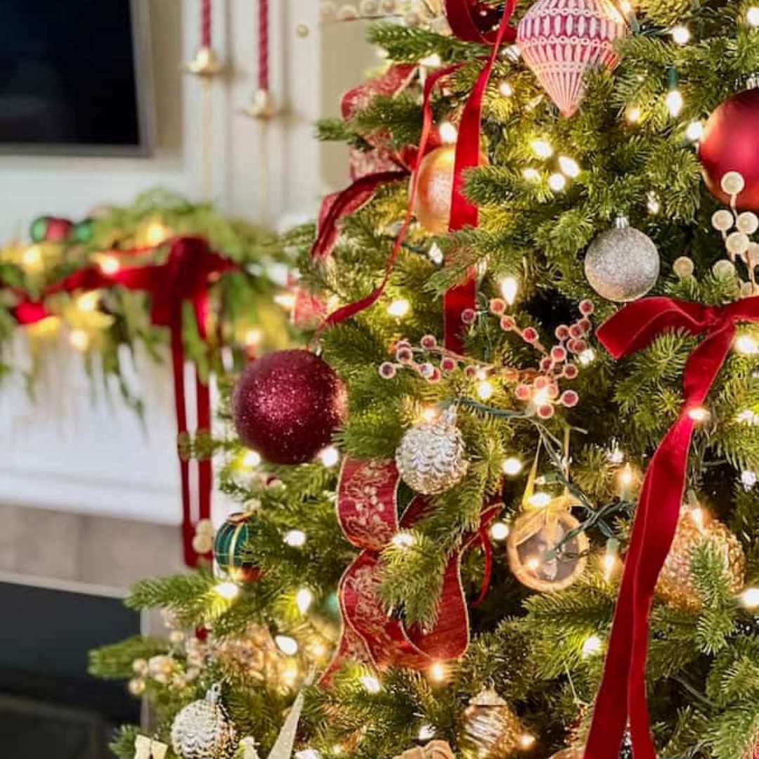 A beautifully decorated Christmas tree adorned with red and gold ornaments, ribbons, and twinkling lights captures the essence of classic Christmas home deco. In the background, a festive garland with red bows is gracefully draped over a fireplace mantel.