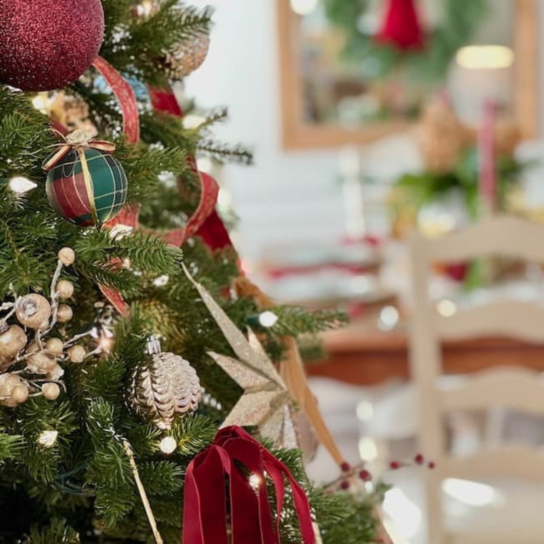 A close-up of a decorated Christmas tree with ornaments, including a plaid ornament, pinecones, a red bauble, and ribbons. The background features Seven on Saturday: a blurred dining table set for the holidays.