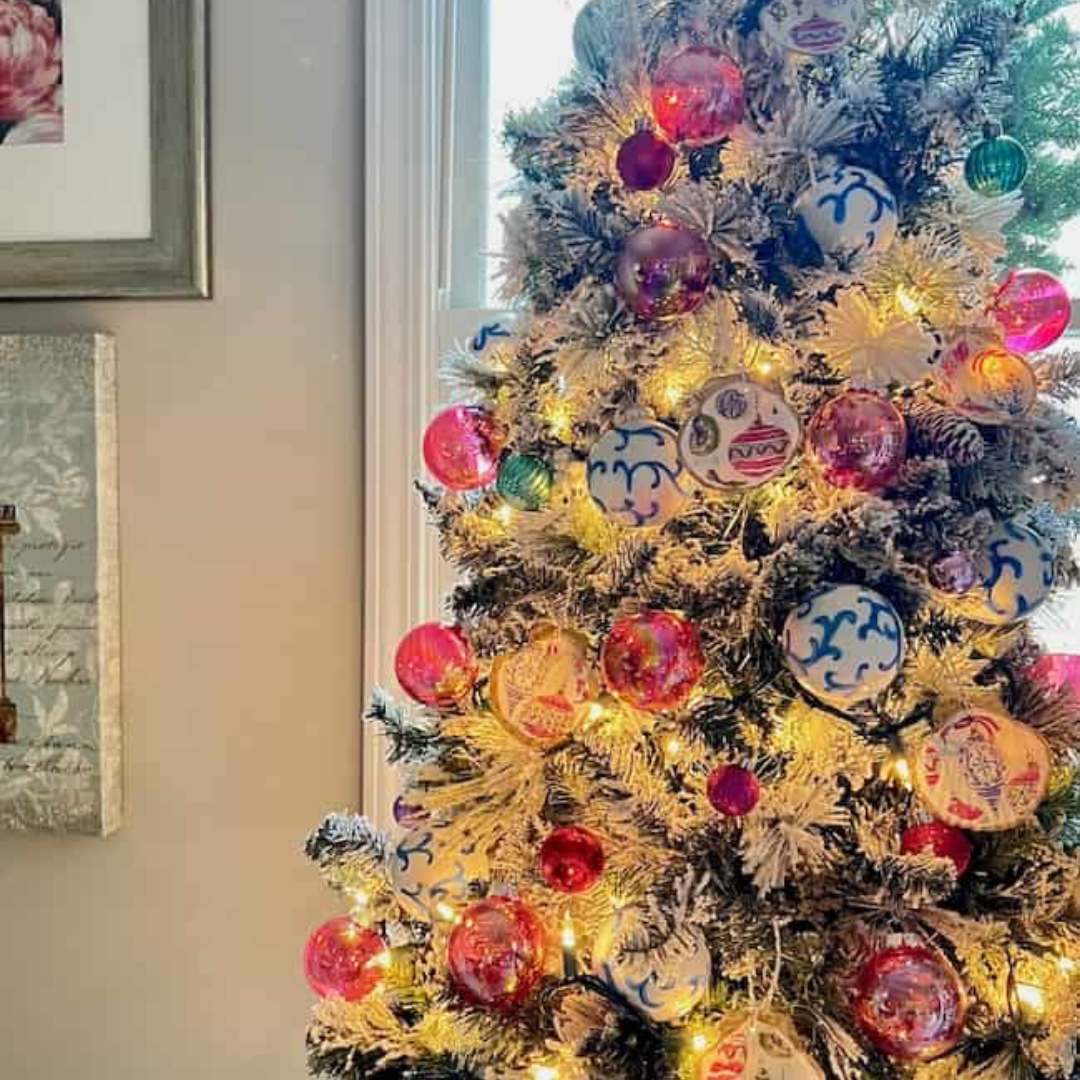 A decorated Christmas tree with frosted branches is adorned with pink and blue ornaments, including charming seashell Christmas ornaments. Warm fairy lights brighten the tree, positioned by a window where natural light spills in. A framed painting hangs on the adjacent wall.