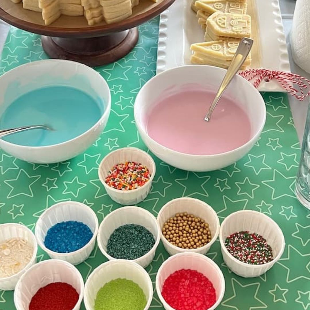 A DIY cookie decorating party awaits, featuring a station with two bowls of colored icing—one blue and one pink—surrounded by small bowls of vibrant sprinkles. In the background sits a tray of undecorated cookies atop a star-patterned tablecloth.