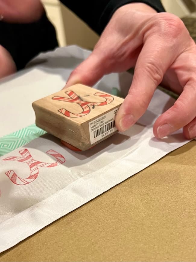 Hands are using a rubber stamp with a candy cane design on a white fabric. The fabric has some red print already on it. A piece of green patterned tape is visible nearby.