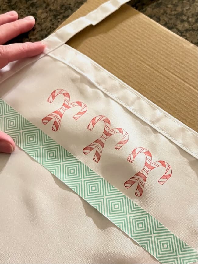 A close-up of a white cloth apron featuring three red and white candy canes above a strip of green geometric pattern fabric. One hand is touching the apron, which is laid on a cardboard surface.