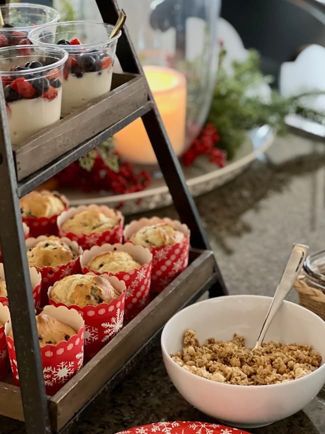 A tiered stand with yogurt parfaits topped with berries and muffins in festive red and white snowflake wrappers. A white bowl filled with granola is in front, and a lit candle is in the background with holiday decor.