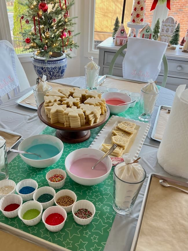 A festive table setup for decorating cookies. Various bowls with colorful icing and sprinkles surround a platter of star-shaped cookies. A small Christmas tree is in the background, adding to the holiday atmosphere.